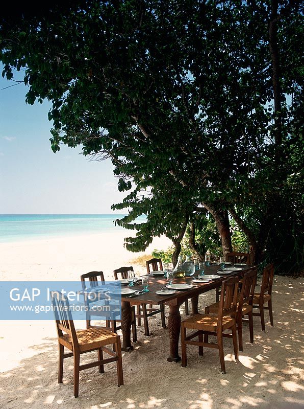 Table à manger et chaises installées sur une plage sous une canopée d'arbres