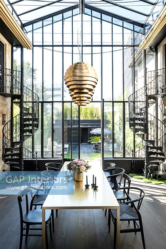 Salle à manger contemporaine dans un bâtiment vitré avec escaliers en colimaçon métalliques des deux côtés et vue sur le jardin