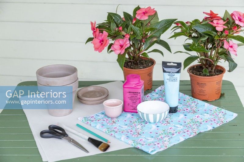 Tissu, pots en terre cuite, soucoupes en terre cuite, peinture acrylique, colle, ciseaux, pinceau, crayon, papier et fleurs disposés sur une table