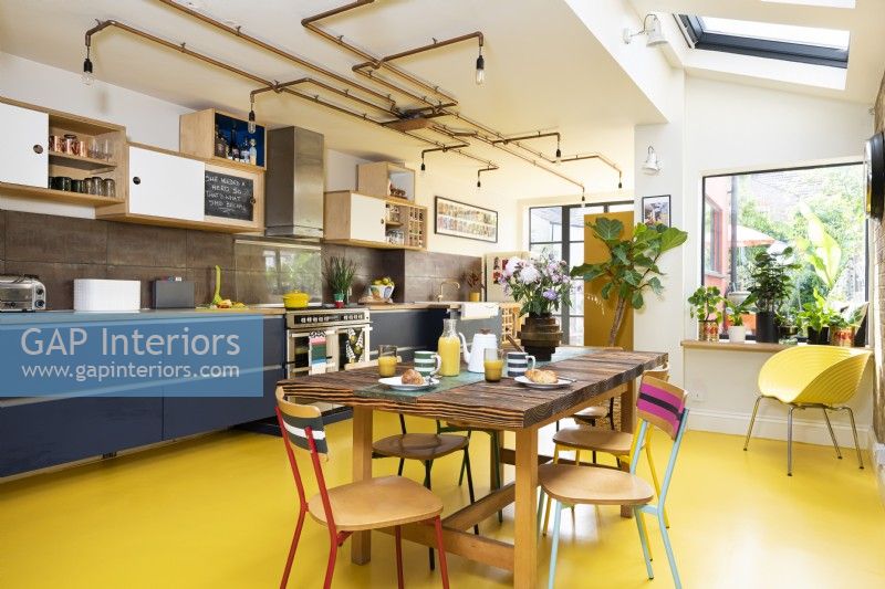 Cuisine rétro moderne avec briques apparentes, sol en caoutchouc jaune, armoires bleues, contreplaqué, éclairage en tuyaux de cuivre et table et chaises recyclées