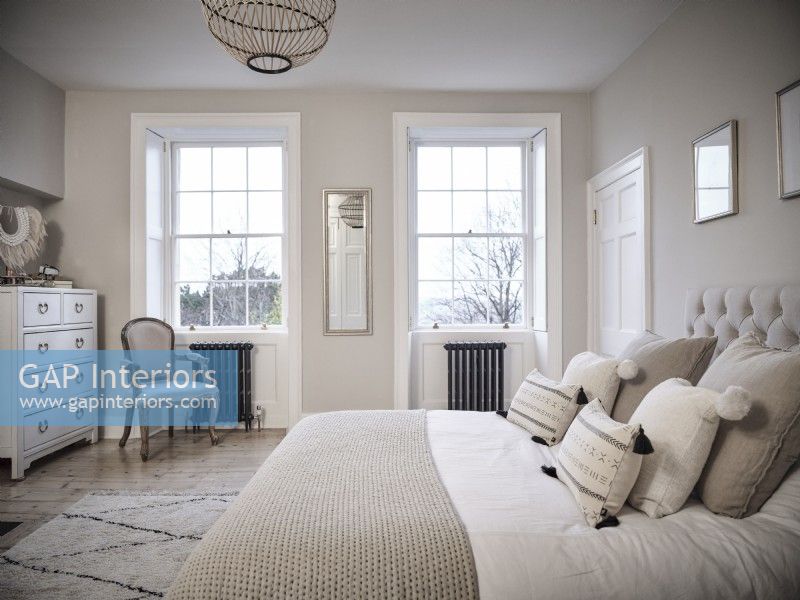 Chambre neutre avec un lit, une chaise rembourrée, des tiroirs et des fenêtres symétriques