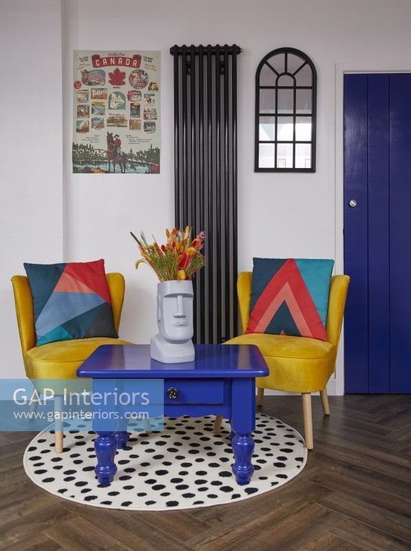 Coin salon avec des chaises jaunes, une table basse bleue et un tapis rond à pois.