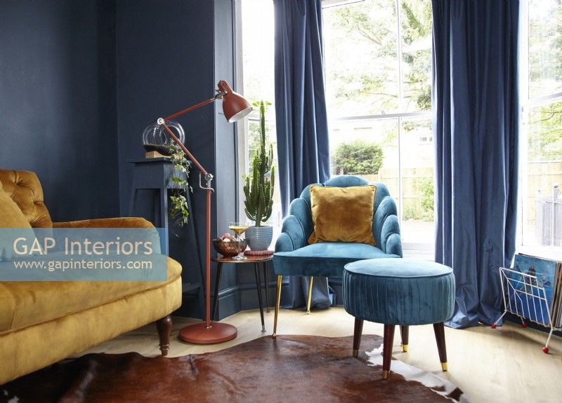 Salon comprenant un canapé jaune moutarde, un fauteuil turquoise avec repose-pieds, un mur peint en bleu marine et un lampadaire.
