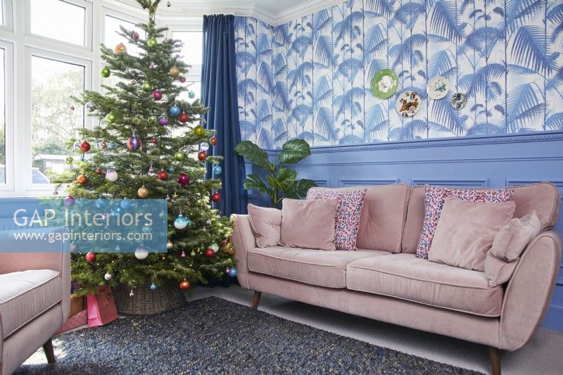 Salon de Noël avec sapin décoré, canapé rose, papier peint imprimé jungle et boiseries bleues.