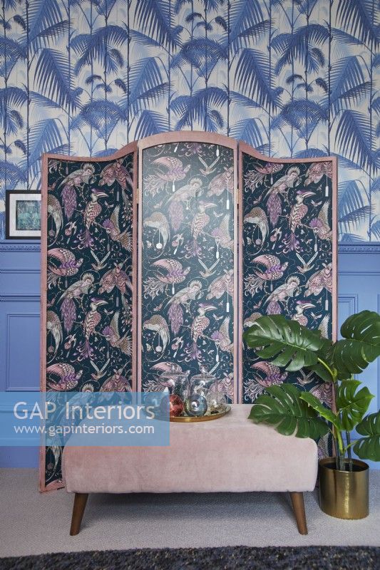 Détail du salon montrant un paravent à motifs, un papier peint imprimé jungle et des lambris bleus.