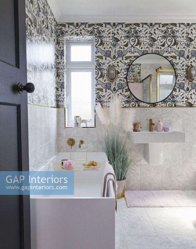 Salle de bain avec papier peint à motifs, miroir rond et robinetterie dorée.