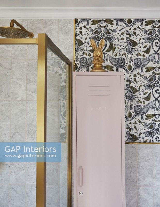 Détail de la salle de bain avec papier peint à motifs, cabine de douche à cadre doré et casier de rangement rose.