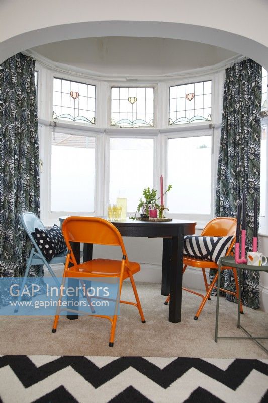 Coin salle à manger avec chaises orange et bleues dans une baie vitrée.