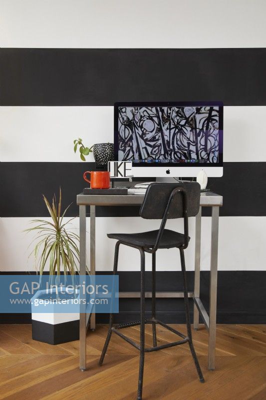 Espace bureau à domicile dans un espace de vie à aire ouverte. Avec des murs rayés noirs et blancs, un tabouret et des plantes.