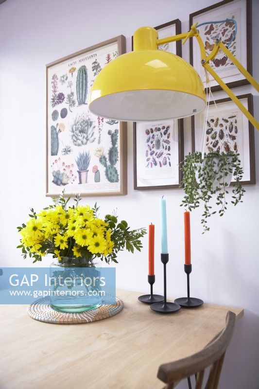 Détail de la table à manger montrant un vase de fleurs, des bougies, un lampadaire jaune avec des imprimés botaniques sur le mur.