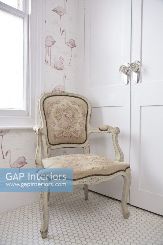 Détail de la salle de bain montrant une chaise ancienne, un papier peint flamant rose et un sol hexagonal blanc.