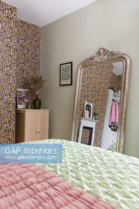 Un grand miroir orné d'un cadre doré au pied d'un lit s'appuie contre un mur vert pâle dans une chambre à coucher avec un papier peint décoré d'un imprimé léopard.