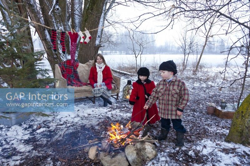 Mère regardant de petits enfants rôtir des guimauves sur un feu ouvert à l'extérieur le jour de Noël. Chaussettes accrochées à la corde après la soirée de patinage