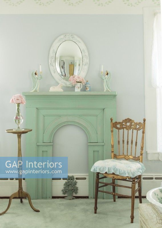 Un miroir convexe est suspendu au-dessus d'une cheminée peinte en vert vieilli. Une chaise et une table dorées insufflent une touche de glamour délavé.
