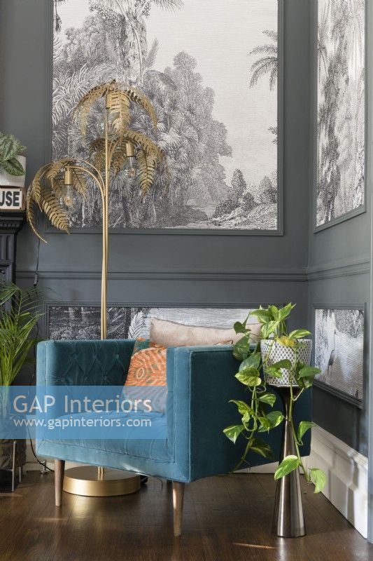 Lampadaire de fougère dorée et une chaise rembourrée bleu sarcelle dans le coin d'un salon peint en gris avec des lambris remplis de papier peint monochrome d'arbres tropicaux