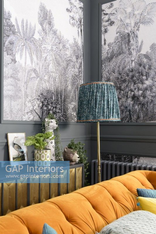 Lampe vintage et meuble de rangement derrière un canapé en velours orange devant des panneaux de papier peint à motifs d'arbres tropicaux noirs et blancs