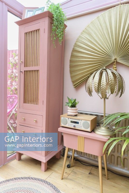 Grande armoire rose recyclée et table de couture vintage peinte en rose récupérée dans une véranda rose avec lampe de table en fougère tropicale dorée