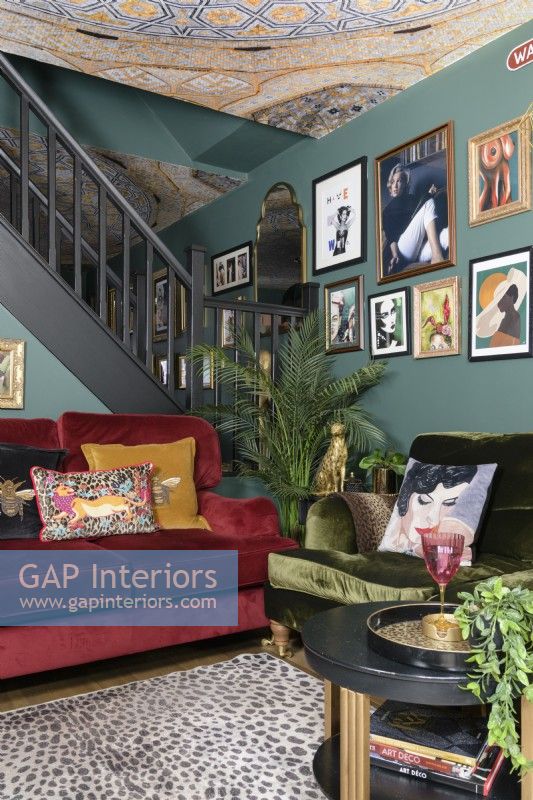 Salon vert ouvert avec des canapés en velours rouge et vert devant un escalier ouvert et un mur d'exposition d'art de style salon et un plafond à motifs en mosaïque