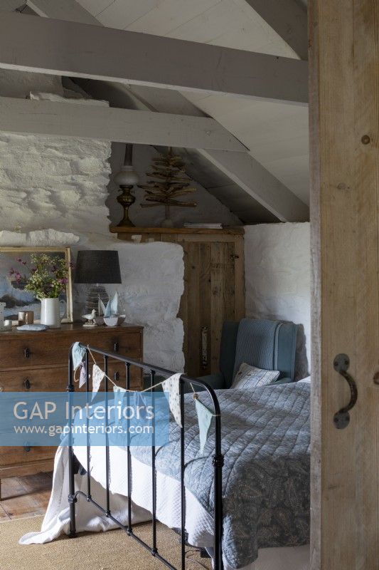 Chambre confortable dans un chalet rustique, murs et poutres blanchis à la chaux, et lit à ossature métallique recouvert d'une couverture bleue confortable