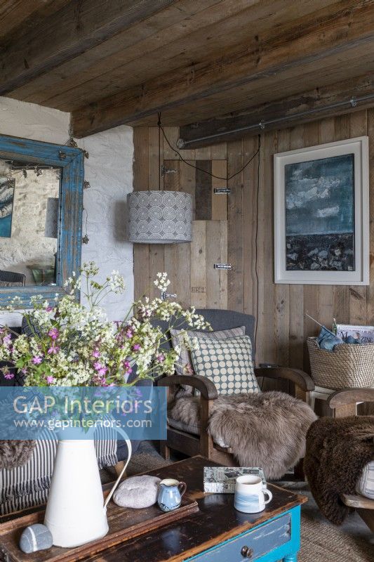 Salon avec murs rustiques, poutres en bois et mobilier confortable. Grand vase de fleurs sauvages cueillies dans la campagne environnante.