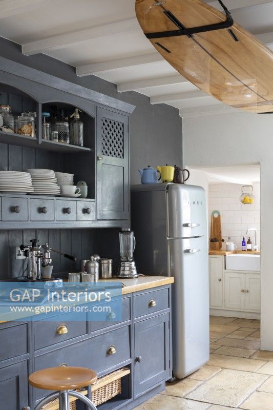 Maison de Sally et John Biddle à Cornwall, armoires de cuisine peintes en bleu et réfrigérateur Smeg moderne, avec planche de surf fixée aux poutres du plafond