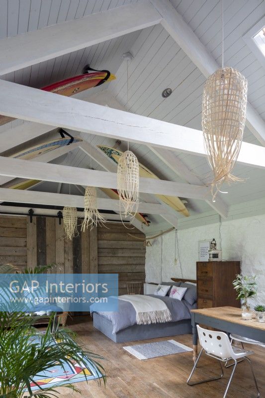 Grange aménagée, avec lit, meubles et bureau. Poutres en bois et décoration rustique, planches de surf décorant le toit en bois