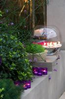 Lit surélevé rendu à côté de la cheminée avec Liriope muscari, des bougies et un grand bol en verre avec des gerberas dans le jardin la nuit
