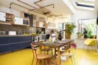 Cuisine rétro moderne avec briques apparentes, sol en caoutchouc jaune, armoires bleues, contreplaqué, éclairage en tuyaux de cuivre et table et chaises recyclées