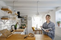 Kati Scalet se tient dans sa cuisine contemporaine en forme de L avec des carreaux de métro blancs et une surface de travail en bois. Sous la fenêtre se trouve une banquette en forme de L.