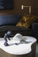 Figurine et sculpture exposées sur une table basse en marbre blanc et laiton devant un canapé bleu foncé et un oreiller doré.