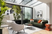 Véranda d'extension moderne avec portes coulissantes en verre, canapé en tissu vert, table d'appoint orange et coussins.