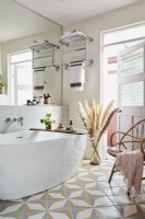 Salle de bain avec baignoire ovale et carrelage au sol, porte miroir murale rose, fauteuil en osier