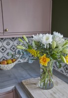 Détail de la cuisine montrant un plan de travail avec un vase de fleurs, des armoires roses et un carrelage à motifs.