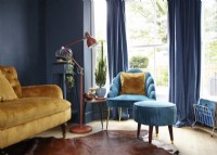 Salon comprenant un canapé jaune moutarde, un fauteuil turquoise avec repose-pieds, un mur peint en bleu marine et un lampadaire.