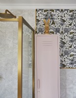 Détail de la salle de bain avec papier peint à motifs, cabine de douche à cadre doré et casier de rangement rose.