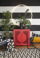 Détail du salon montrant une armoire rouge moderne, une lumière circulaire, des chaises en métal contemporaines et des plantes. Avec des murs peints à rayures noires et blanches et un tapis à motifs.