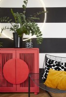 Détail du salon montrant une armoire rouge moderne, une lumière circulaire, des chaises en métal contemporaines et des plantes. Avec des murs peints à rayures noires et blanches et un tapis à motifs.
