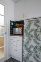 Détail de la cuisine montrant un réfrigérateur recouvert de vinyle jungle, un four micro-ondes et une corbeille de fruits.