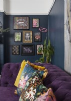 Détail du salon montrant des murs bleu foncé avec des œuvres d'art et un canapé violet avec des coussins imprimés.