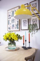 Détail de la table à manger montrant un vase de fleurs, des bougies, un lampadaire jaune avec des imprimés botaniques sur le mur.