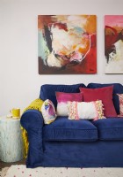 Détail coloré du salon avec un canapé en velours bleu foncé, des coussins colorés et de grands tableaux abstraits.