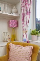 Détail du salon montrant un fauteuil jaune avec une lampe de table et des rideaux roses.