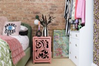 Un mur en briques apparentes dans une chambre avec une table de chevet en rotin peint en rose à côté d'un lit en velours vert et une armoire ouverte à emporter et un meuble à tiroirs.