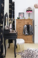 Tabouret recouvert de fourrure blanche et une coiffeuse décorative recyclée peinte en noir avec un plateau recouvert d'un imprimé léopard en vinyle devant une commode en pin