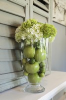 Un vase transparent contient un arrangement simple d'hortensias, de pommes et de citrons verts dans la même famille de couleurs qui fait entrer le jardin.