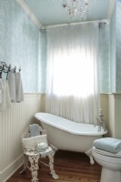 L'association d'une baignoire à pantoufle vintage élégante avec une chaise de jardin en fonte et un papier peint à motifs à volutes avec un panneau de lambris apporte une élégance de cottage classique à la salle de bain d'invités.