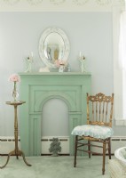 Un miroir convexe est suspendu au-dessus d'une cheminée peinte en vert vieilli. Une chaise et une table dorées insufflent une touche de glamour délavé.