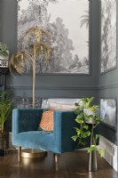 Lampadaire de fougère dorée et une chaise rembourrée bleu sarcelle dans le coin d'un salon peint en gris avec des lambris remplis de papier peint monochrome d'arbres tropicaux