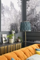 Lampe vintage et meuble de rangement derrière un canapé en velours orange devant des panneaux de papier peint à motifs d'arbres tropicaux noirs et blancs