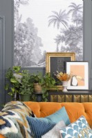 Détail derrière un canapé en velours orange d'une étagère avec des photos encadrées et des plantes d'intérieur devant un panneau de papier peint à motifs d'arbres tropicaux noir et blanc